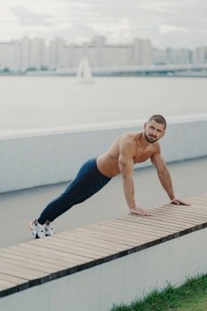 Muscular man warms up outdoors, planks with naked torso, enjoys open-air exercise, wears activewear for vitality.