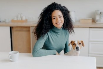 Curly-haired woman in turtleneck sits at kitchen table, enjoys tea, plays with Jack Russell terrier. Joyful moment with beloved pet.