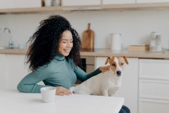 Joyful Afro woman bonds with her dog, laughs in cozy kitchen, sips coffee, cherishes the domestic bliss. Pet-loving happiness.
