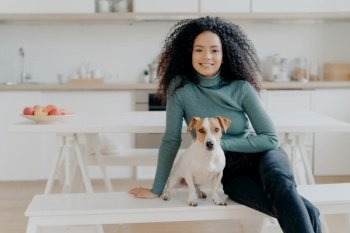 Happy Afro woman sits with dog, poses in kitchen, expresses joy, relaxes with beloved pet. Loving home companionship.