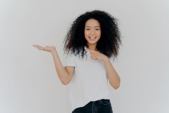 Confident girl with Afro hair points and smiles, chooses, wears casual clothes, poses on white background.