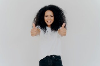 Cheerful Afro-haired woman gives thumbs up, approves with a smile, indoors, isolated on white background.