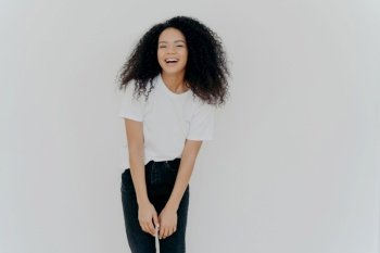 Joyful African American woman in casual wear laughs happily, poses against white background with blank space, feeling energized.