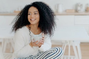 Cheerful woman with curly hair smiles, holds coffee mug, enjoys cozy morning in kitchen. Afro lady on day off.