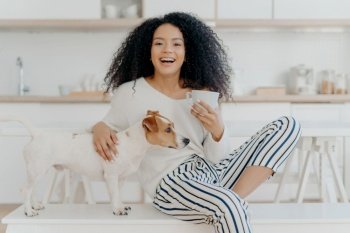 Joyful woman with dog in kitchen, petting her favorite Jack Russell Terrier, enjoying hot beverage, and smiling happily.