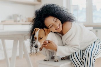 Lovely curly woman embraces beloved dog with care, wearing stylish clothes, posing at home, expressing affection.
