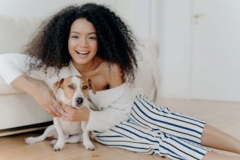Happy curly woman with fashionable clothes plays with a funny little dog on the floor, sharing joyful moments and love.