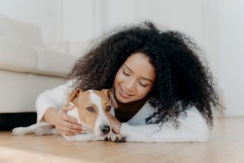 Relaxed Afro woman in white sweater plays and pets a cute puppy, enjoying the company of her beloved Jack Russell terrier.