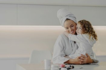 Loving mom embraces daughter after spa treatments, wearing white bathrobes, in modern kitchen at home.