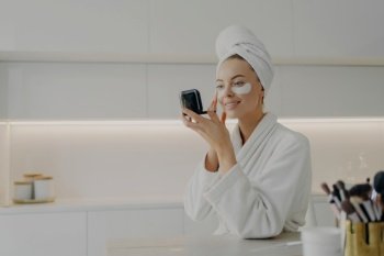 Woman in bathrobe applies under-eye patches, checks mirror in kitchen during morning skincare routine.