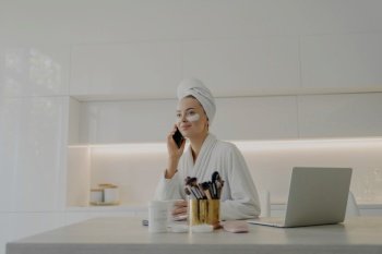 Busy young woman in bathrobe multitasks, talks on phone, works on laptop, wears under-eye patches, prepares for work at home.