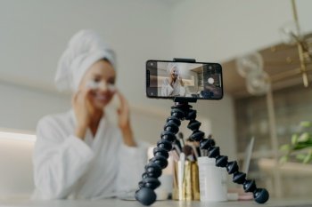 Beauty blogger records under-eye patch tutorial, tackles wrinkles and dark circles, focused on smartphone, bathrobe attire.