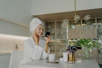 Woman in bathrobe applies face cream, checks reflection in compact mirror. Skincare routine in kitchen.
