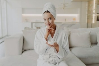 Woman in bathrobe with cream jar, smiles at camera, sits on sofa, does cosmetic procedures, skincare concept.