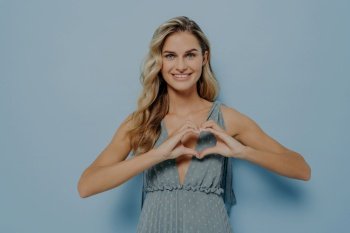 Blonde woman smiling, showing heart gesture, expressing love and affection, isolated on blue background.