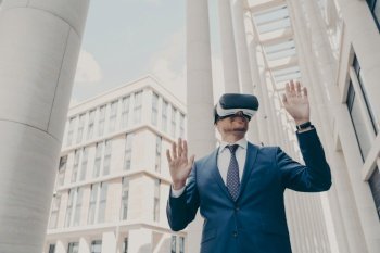 Happy businessman in VR goggles outdoors, managing project in cyberspace, testing futuristic VR headset, blurred buildings in background.
