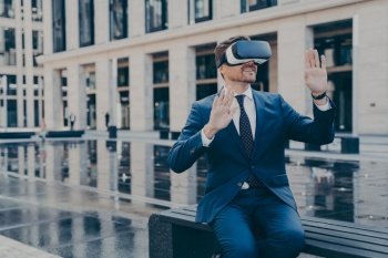 Man in VR goggles reaching in virtual reality, sitting on bench near fountain and office buildings during coffee break.