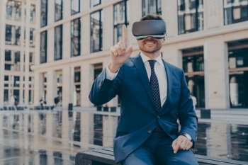 Happy businessman in blue suit testing VR goggles on bench, controlling with finger. Excited man outdoors, office buildings blurred.