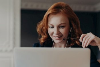 Focused freelancer watches tutorial webinar, uses earphones, during online seminar. Happy woman entrepreneur in close-up at laptop.