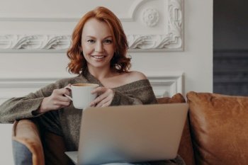 Ginger female on sofa with coffee searches job online, prepares presentation, enjoys cozy atmosphere, high-speed internet.
