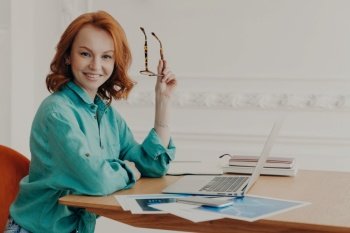 Ginger woman in coworking space awaits online meeting, holding glasses, smiling at camera, working from home.