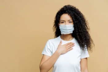 Serious Afro American woman, medical face mask, breathing difficulty, hand on chest, infected with COVID-19. Isolated, beige background. Health care concept.