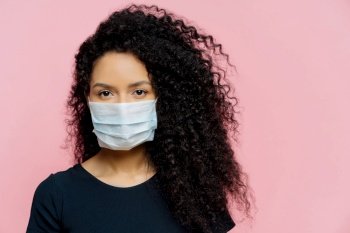 Serious dark-skinned woman on home isolation. Wearing medical mask, casual attire. Quarantine at home. Pink background. COVID-19.