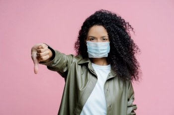 Curly-haired woman wearing medical mask, thumbs down gesture. Health-conscious, virus prevention concept.