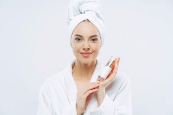 Calm woman with healthy skin holds body lotion, poses in bath robe after shower, advertises product. 