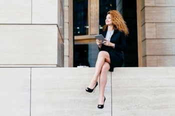 Elegant woman in black suit, high heels, slender legs. Happy expression, sits with tablet, looks aside. Office worker poses outdoors.