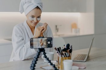 Pleased beauty blogger shares skin care experience, wears eye patches, poses at table with tools, in bathrobe with towel.