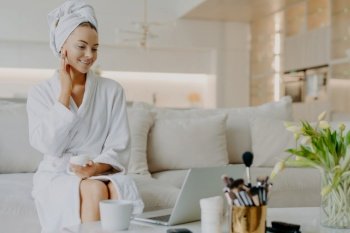 Smiling woman applies cream, advises skincare, in robe, towel, on sofa, rejuvenation procedure, laptop screen. Cosmetology.