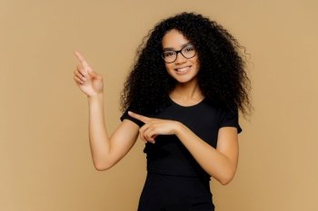 Happy African American woman points up and aside, demonstrates blank copy space, wears spectacles, casual t-shirt, gentle smile. Promo perfection.