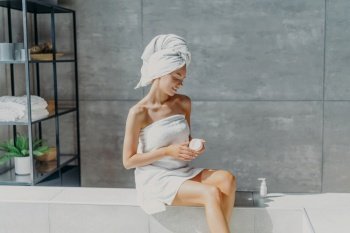Relaxed European woman applies leg cream, wrapped in towel. Beauty treatment, healthy skin. Hygiene concept.