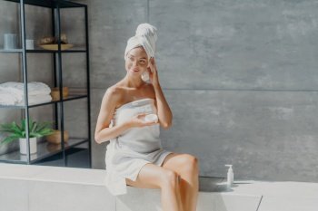 Dreamy European woman applies body cream, wrapped in towel. Health, beauty concept. Bathroom pampering.