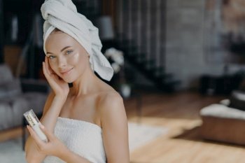 Skin care concept. Woman applies face lotion, wrapped in towel. Daily pampering, facial treatments. Bare shoulders, healthy complexion.