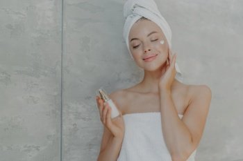 Woman applies face cream, holds lotion bottle, enjoys skin pampering. Towel on head, grey wall background.