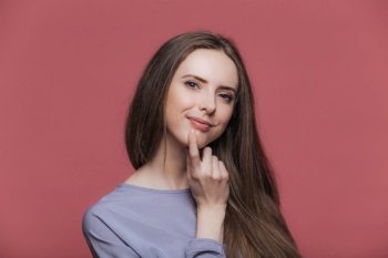 Pleased attractive female with luxurious brunette hair, hand under chin, looks delightfully at camera. Isolated on pink background. Positive emotions concept.