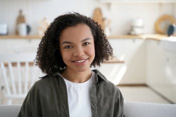 Friendly mixed-race girl with afro hair, smiling at home. Happy teen on sofa, enjoying sunny lazy weekend in cozy house.