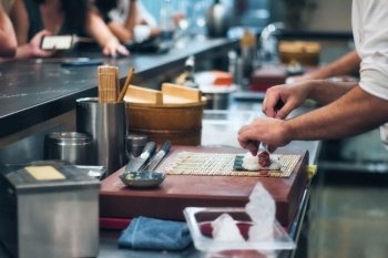 Close-up of a chef’s hands rolling a sushi roll with rice and seaweed at a sushi bar with people in the background at the counter