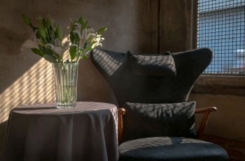A Wooden Upholstered Chair and the Bouquet of Flowers in a Glass Vase on Table in the Room with Bare Cement Wall. They are affected by the Sunbeam Shining through Steel Mesh Window in the Afternoon. Space for text, Selective focus.