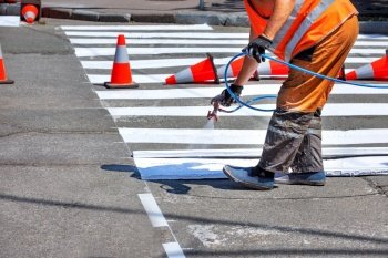 A road worker applies white road markings to a crosswalk using an airbrush and a wooden template surrounded by traffic cones.. A road worker in orange overalls airbrushes white road markings at a pedestrian crossing.