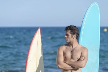 A Hispanic surfer guy standing on the beach in a swimsuit looking at the camera next to two surfboards.