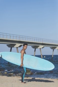 Hispanic surfer boy standing on the beach in neoprene holding his blue surfboard at sunset