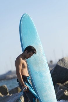 Hispanic surfer boy standing on the beach in neoprene holding his blue surfboard at sunset