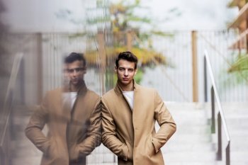 Portrait of stylish young man with coat standing outdoors and leaning on wall.