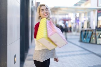Blonde smiling woman holding shopping bags while standing next to a shopping mall