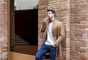 Front view of fashionable young man wearing denim clothes leaning on a wall while using a mobile phone