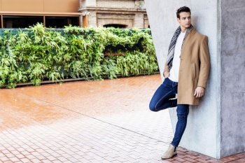 Young man wearing winter clothes in the street. Young guy with modern hairstyle with coat, blue jeans and white sweeter.
