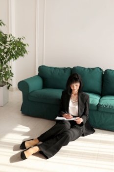 smiling businesswoman writes in note pad and looking in camera. brunette woman is sitting close to green sofa in a black suit and high heels shoes. Green houseplant in interior. Work from home. smiling businesswoman writes in note pad and looking in camera. brunette woman is sitting close to green sofa in a black suit and high heels shoes. Green houseplant in interior. Work from home.